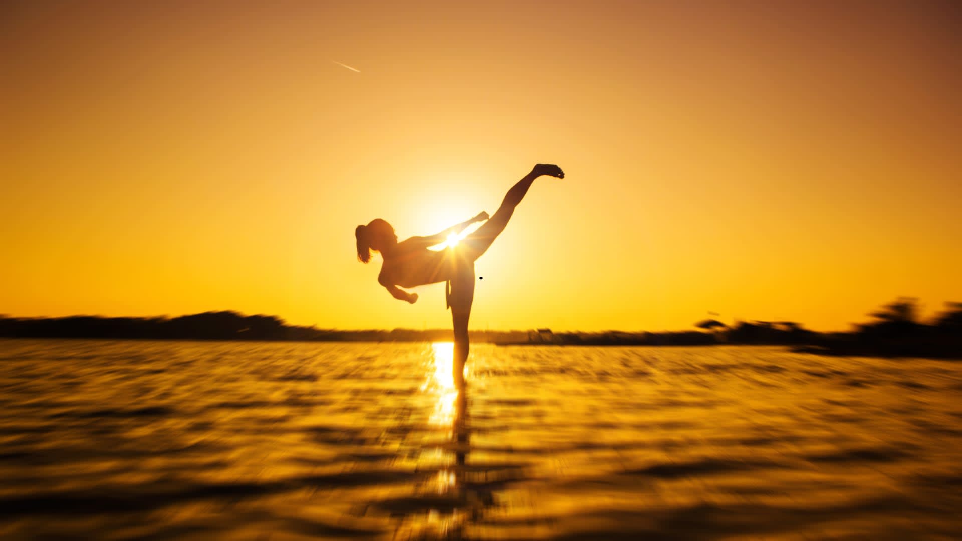 Silhouette of a martial artist holding a high kick in the water at sunset